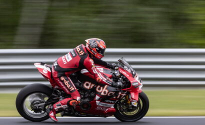 MotoGP rider in red livery leaning into a high-speed turn on a race track, blurred background behind metal barriers.