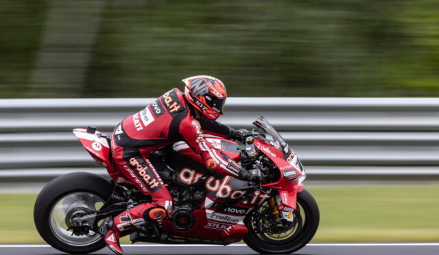 Racer in red suit riding a red sport motorcycle on a track with guard rails in the background, motion blur indicating speed.