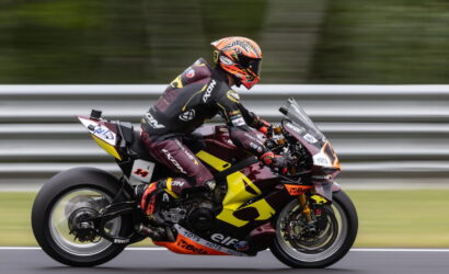Motorcycle racer in full gear leans into a high-speed corner on a yellow-and-maroon racing bike on a track, gravel and barriers blurred in the background