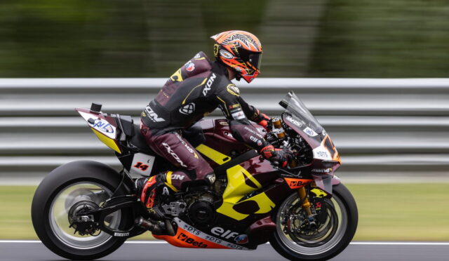 Motorcycle racer in full gear leans into a high-speed corner on a yellow-and-maroon racing bike on a track, gravel and barriers blurred in the background