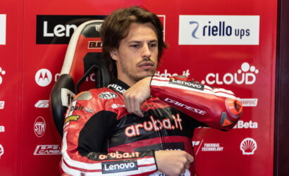 Motorcycle racer in red-and-black suit sits in a chair against a red sponsor wall, arms crossed over chest looking thoughtful.