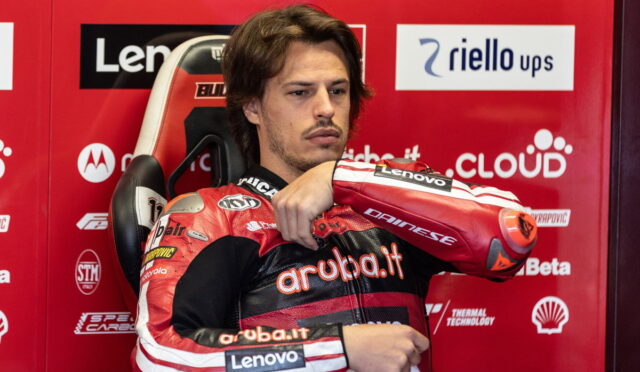 Motorcycle racer in red-and-black suit sits in a chair against a red sponsor wall, arms crossed over chest looking thoughtful.