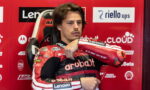 Moto racer in red/black leather suit sits in a racing chair, arm raised as he adjusts his suit against a red sponsor-filled backdrop)