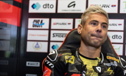 Moto racer in a black-yellow racing suit sits in a chair against a sponsor-backed backdrop, looking to the side.”] ,