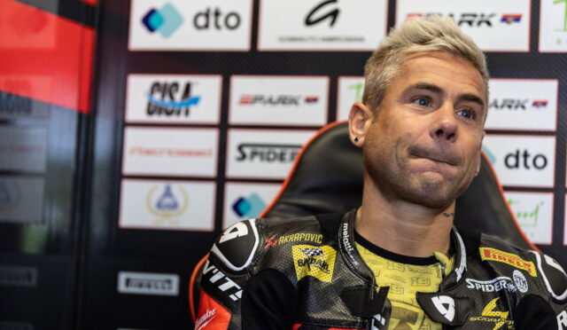 Moto racer in a black-yellow racing suit sits in a chair against a sponsor-backed backdrop, looking to the side.”] ,
