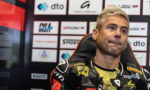 Racer in black-and-yellow leather suit sits in a chair before a wall of sponsor logos during a press event, looking slightly upward as if listening or thinking.