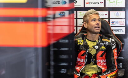 Racer in a red-and-black racing suit sits in a padded chair against a sponsor-filled backdrop during a pit interview or press event.