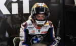 Moto racing driver in white, blue, and black suit sits in the pit area with helmet on, ready for the race