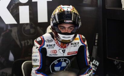 Moto racing driver in white, blue, and black suit sits in the pit area with helmet on, ready for the race