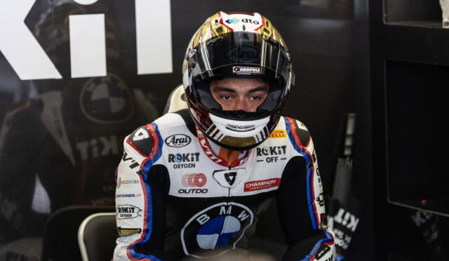 Moto racing driver in white, blue, and black suit sits in the pit area with helmet on, ready for the race
