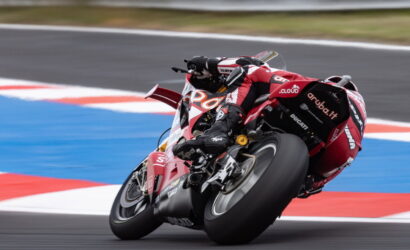 Red Ducati racing motorcycle with a rider in matching red leathers leaning into a sharp track corner, blue runoff visible on the apron.