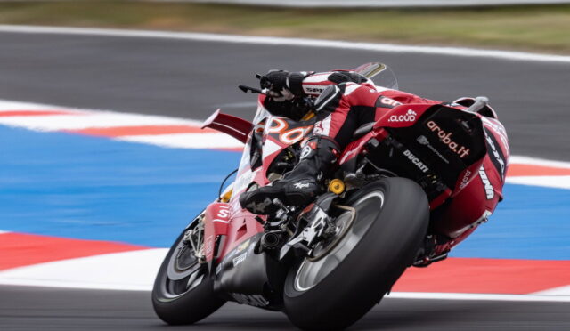 Red Ducati racing motorcycle with a rider in matching red leathers leaning into a sharp track corner, blue runoff visible on the apron.