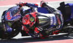 Rider in a red helmet leaning into a turn on a blue Yamaha race motorcycle on the track, with sponsor logos visible.