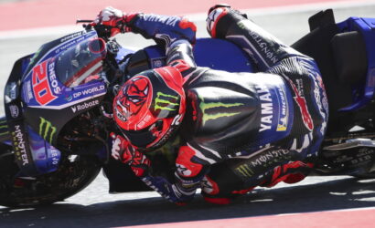Rider in a red helmet leaning into a turn on a blue Yamaha race motorcycle on the track, with sponsor logos visible.