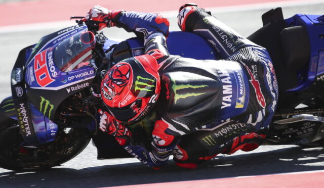 Rider in a red helmet leaning into a turn on a blue Yamaha race motorcycle on the track, with sponsor logos visible.