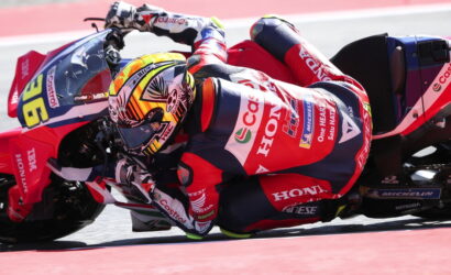 Motorcycle racer in a red Honda racing suit leaning into a sharp turn on a racetrack at speed.