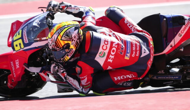 Motorcycle racer in a red Honda racing suit leaning into a sharp turn on a racetrack at speed.