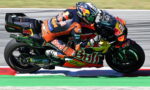 Professional motorcycle racer in orange-and-blue Red Bull gear leaning into a corner on a race track, sport bike adorned with sponsor logos.