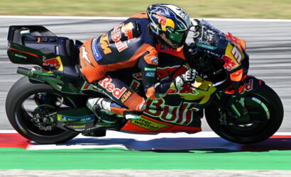 Professional motorcycle racer in orange-and-blue Red Bull gear leaning into a corner on a race track, sport bike adorned with sponsor logos.