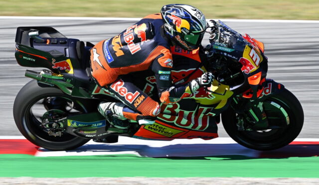 Professional motorcycle racer in orange-and-blue Red Bull gear leaning into a corner on a race track, sport bike adorned with sponsor logos.