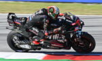 Two riders on a high-performance racing motorcycle lean into a left-hand corner on a track, wearing black and red branded leathers with sponsor logos in a tight slipstream close to the curb.