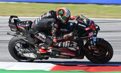Two riders on a high-performance racing motorcycle lean into a left-hand corner on a track, wearing black and red branded leathers with sponsor logos in a tight slipstream close to the curb.