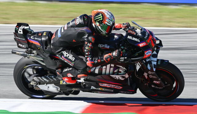 Two riders on a high-performance racing motorcycle lean into a left-hand corner on a track, wearing black and red branded leathers with sponsor logos in a tight slipstream close to the curb.