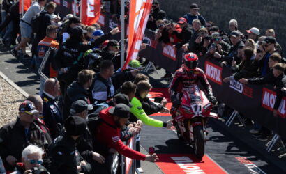 Motorcycle racer in red suit rides a red bike toward the finish barrier as crowd cheers behind barriers lined with Motul banners.