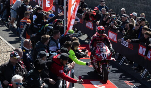 Motorcycle racer in red suit rides a red bike toward the finish barrier as crowd cheers behind barriers lined with Motul banners.