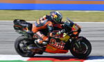 MotoGP rider in an orange racing suit on a Red Bull KTM bike leaning into a turn on the racetrack.