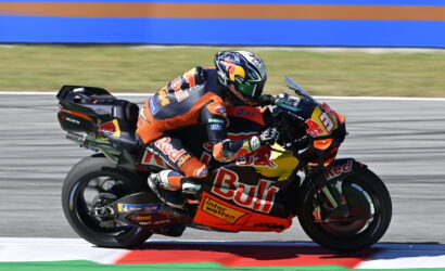 MotoGP rider in an orange racing suit on a Red Bull KTM bike leaning into a turn on the racetrack.