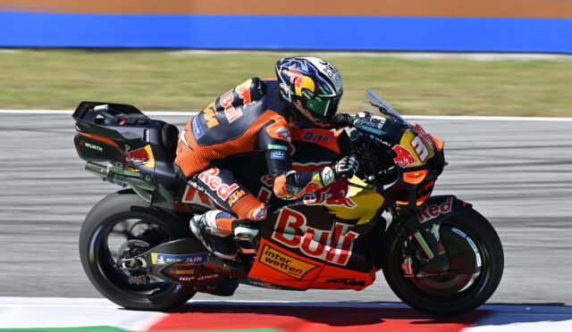 MotoGP rider in an orange racing suit on a Red Bull KTM bike leaning into a turn on the racetrack.
