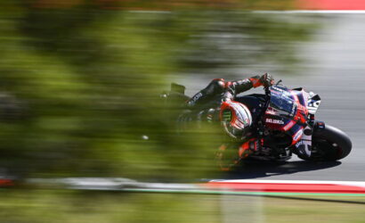 Motorcycle racer in red-and-black livery leans into a turn on a race track, with blurred foreground greenery suggesting speed.