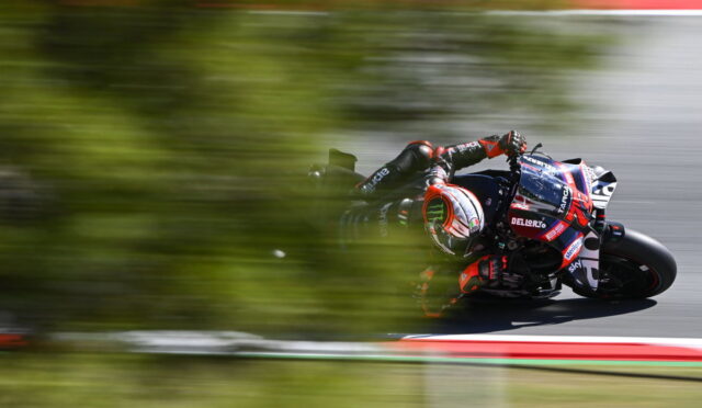 Motorcycle racer in red-and-black livery leans into a turn on a race track, with blurred foreground greenery suggesting speed.