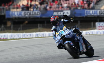 Motorcycle racer in blue gear leans into a sharp turn on a race track, with spectators in the stands watching above the track barriers.