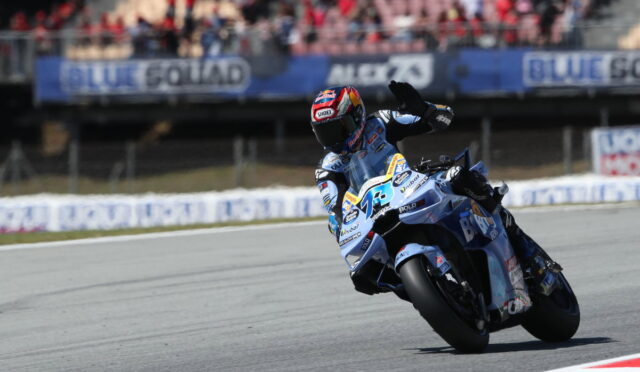 Motorcycle racer in blue gear leans into a sharp turn on a race track, with spectators in the stands watching above the track barriers.