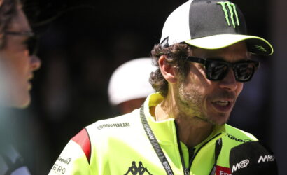 Man in a bright neon jacket being interviewed, wearing sunglasses and a black-and-white cap with a Monster logo, microphone nearby