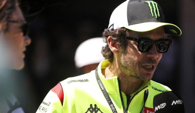 Man in a bright neon jacket being interviewed, wearing sunglasses and a black-and-white cap with a Monster logo, microphone nearby