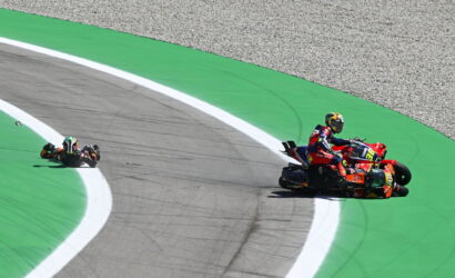 Two motorcycle racers lean into a tight turn on a bright green race track, one bike trailing near the white line on the apex edge.