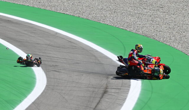 Two motorcycle racers lean into a tight turn on a bright green race track, one bike trailing near the white line on the apex edge.