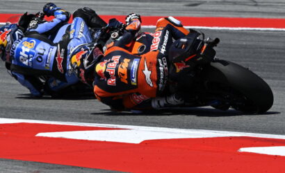 Group of MotoGP riders leaning into a sharp turn on a race track, KTM in orange leading the pack with Red Bull branding.