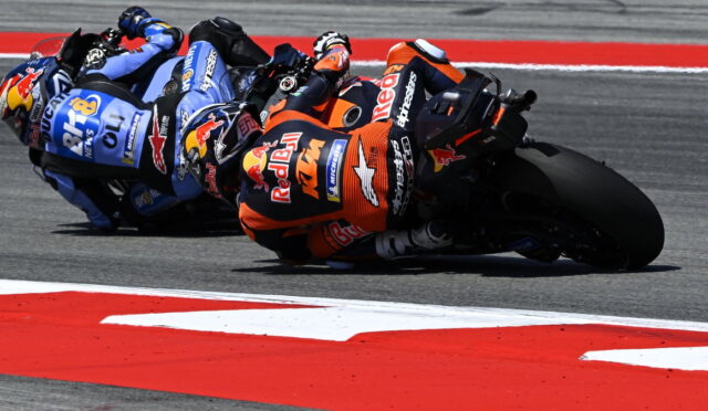 Group of MotoGP riders leaning into a sharp turn on a race track, KTM in orange leading the pack with Red Bull branding.
