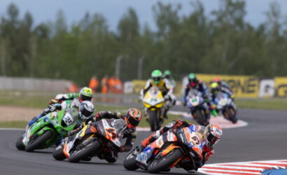 MotoGP-style race: a group of riders leaning into a sharp turn on a racetrack, colorful helmets and bikes in motion with blurred background trees and barriers.