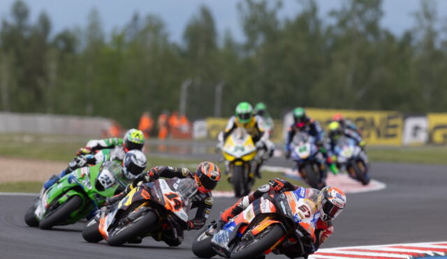 MotoGP-style race: a group of riders leaning into a sharp turn on a racetrack, colorful helmets and bikes in motion with blurred background trees and barriers.