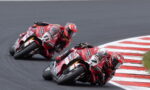 Two red racing motorcycles lean into a sharp corner on a dark asphalt track, riding close to red-and-white curbs.