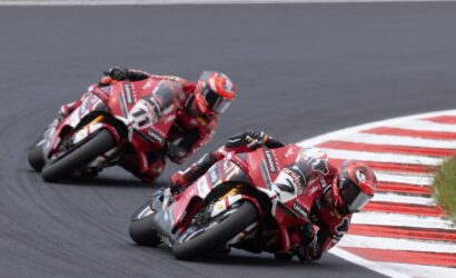 Two red racing motorcycles lean into a sharp corner on a dark asphalt track, riding close to red-and-white curbs.