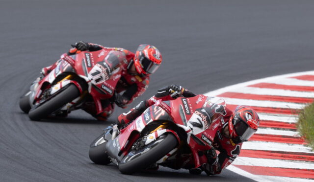 Two red racing motorcycles lean into a sharp corner on a dark asphalt track, riding close to red-and-white curbs.