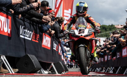 Moto racing rider on a red-and-white sport bike rides toward the front with a cheering crowd and sponsor banners along the barriers, after a race.