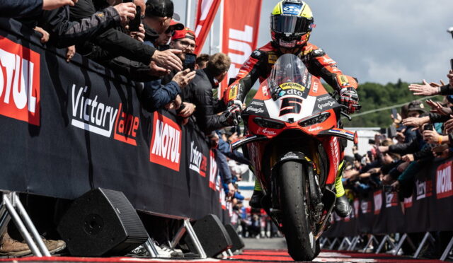 Moto racing rider on a red-and-white sport bike rides toward the front with a cheering crowd and sponsor banners along the barriers, after a race.