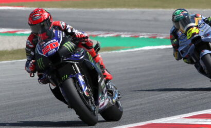 Two motorcycle racers lean into a sharp bend on a race track, the lead rider wearing red helmet and a blue-and-black Yamaha Monster energy bike #20.